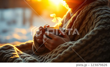 A person bundled in a cozy knit blanket holds a steaming mug, bathed in the warm glow of sunlight, evoking a sense of comfort and tranquility 123879826