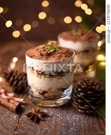 Festive dessert in a glass with layers of cream, chocolate, and crumbs, topped with cocoa powder and a sprig of greenery, surrounded by pinecones and spices 123879890