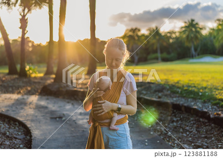 A loving mother walks through a peaceful park, carrying her baby securely in a sling. A heartwarming moment of maternal bonding, babywearing, and the joy of motherhood in nature 123881188