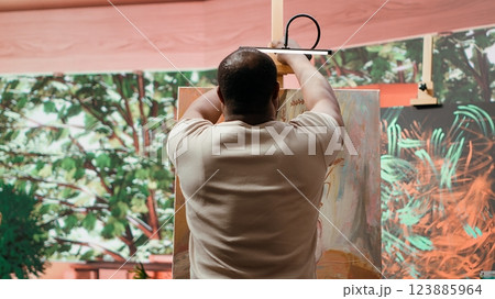 Painter is mixing multiple colors on a palette to apply the perfect shade, creating a beautiful rustic painting on canvas. Male artist using artistic tools and supplies for his hobby. Camera A. 123885964