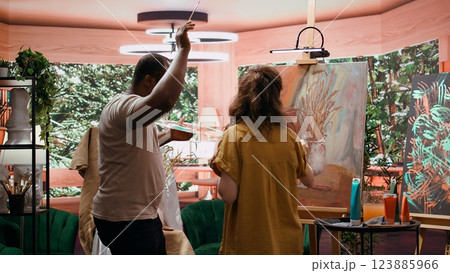 Joyful artist and her apprentice applying watercolor paint on canvas, working together to create a beautiful masterpiece in atelier. Two people painting a colorful scene with music. Camera B. 123885966