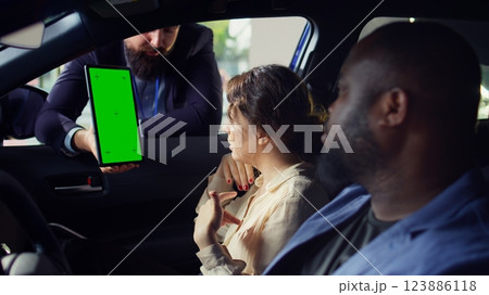 Salesman using green screen tablet to present car addons options to clients, after finishing test drive. Showroom agent mockup device to customers driving vehicle, camera B close up Salesman using green screen tablet to present car addons options to clients, after finishing test drive. Showroom agent mockup device to customers driving vehicle, camera B close up 123886118
