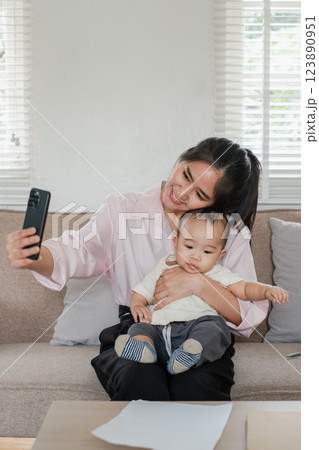 Smiling mother holding her baby while taking a selfie on a comfortable couch in a well-lit living room. 123890951