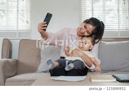 Smiling mother and baby capturing a selfie on a comfortable sofa in a well-lit living room, showcasing family bonding and modern technology. 123890952
