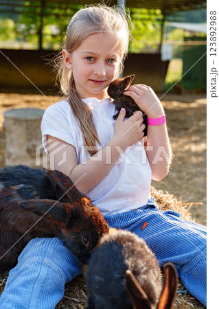 Young girl holds a small rabbit while sitting among playful bunnies at a farm during a sunny afternoon 123892988