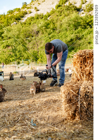 A man films adorable rabbits while kneeling in a sunny farmyard 123892989