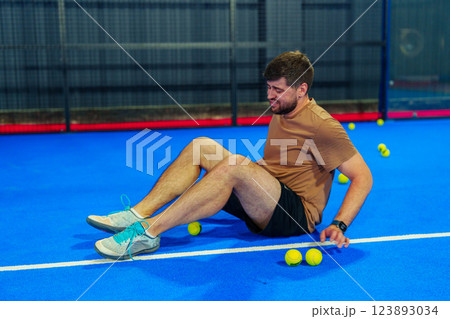 Young man engages in muscle recovery on a blue sports court surrounded by tennis balls 123893034