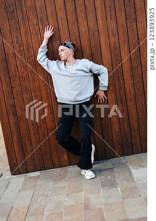 A young man wearing a gray sweater and black pants leans against a wooden wall, looking off to the side 123893925