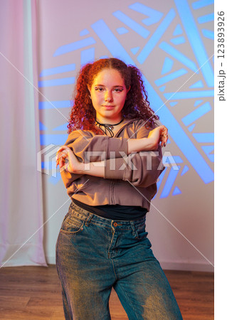 A young woman with curly red hair stands in a studio, posing with her arms crossed. She is wearing a light brown hoodie and blue jeans A young woman with curly red hair stands in a studio, posing with her arms crossed. She is wearing a light brown hoodie and blue jeans 123893926