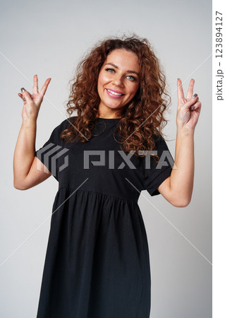 A young woman with curly hair poses cheerfully with peace signs in a neutral setting 123894127