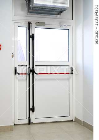 Modern double door with red safety bar in a clean, well-lit hallway of a building Modern double door with red safety bar in a clean, well-lit hallway of a building 123894251