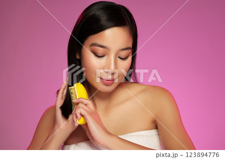 Woman styling her long black hair with a yellow brush against a vibrant pink background during a portrait session 123894776