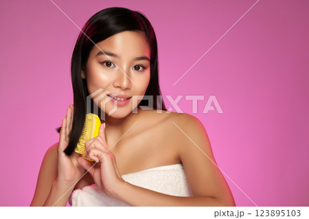 Young woman with long hair holding a yellow comb contrasts against a vibrant pink background while smiling confidently 123895103