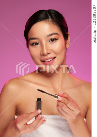 Woman applying mascara while smiling in a beauty setting with soft pink background 123895207