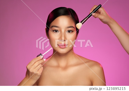 Makeup artist applies lip gloss and blush to model with pink backdrop during studio photoshoot Makeup artist applies lip gloss and blush to model with pink backdrop during studio photoshoot 123895215
