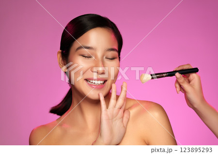 Woman smiling while receiving makeup application in a brightly lit studio with pink backdrop 123895293