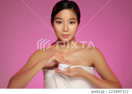 Young woman applying skincare product with towel on her shoulders in a vibrant pink background Young woman applying skincare product with towel on her shoulders in a vibrant pink background 123895322