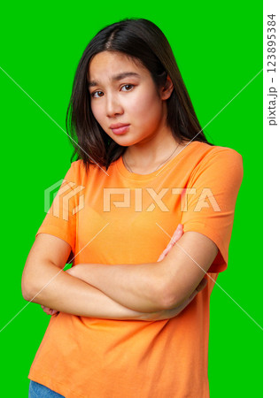 Young woman with crossed arms shows a contemplative expression against a bright green background in a casual orange shirt Young woman with crossed arms shows a contemplative expression against a bright green background in a casual orange shirt 123895384