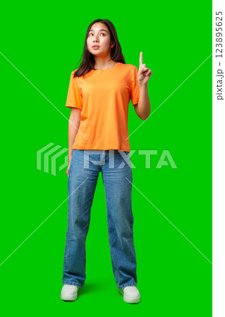 Young woman in bright orange shirt stands with finger raised on green background 123895625