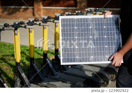 Male person holding solar panel in front of line of electric scooters parked in park. Integration of sustainable renewable energy, showcasing clean energy solution for charging electric scooters. 123902922