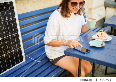 Young woman charging her smartphone from solar panel while relaxing. Pretty girl drinking coffee and checking her phone next to solar battery. Conscious female choosing sustainable lifestyle. 123902931