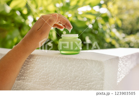 Close-up of a woman hand with white moisturizer on her fingers and a green jar. Cosmetic product with anti-aging cream on the background of bright green foliage. Close-up of a woman hand with white moisturizer on her fingers and a green jar. Cosmetic product with anti-aging cream on the background of bright green foliage. 123903505