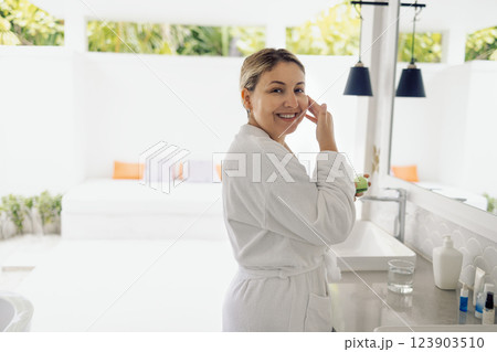A young woman in a white bathrobe applies cream to her neck in the bathroom A young woman in a white bathrobe applies cream to her neck in the bathroom 123903510