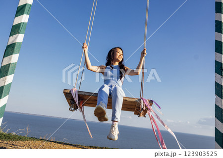 Attractive Asian girl in casual clothes swinging on an outdoor swing. A happy Korean woman laughs and enjoys life. Seascape and blue sky. 123903525