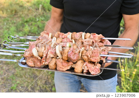 Pieces of raw pork strung on skewers lie on an iron tray held by a man, ready for grilling or cooking. The image highlights the preparation process, perfect for themes of food, cooking, and barbecue. Pieces of raw pork strung on skewers lie on an iron tray held by a man, ready for grilling or cooking. The image highlights the preparation process, perfect for themes of food, cooking, and barbecue. 123907006