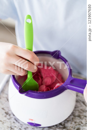 Eager eyes focus on the pot of vibrant, melting candy as a young cook gently swirls the mixture, poised to transform plain snacks into delectable treats. Eager eyes focus on the pot of vibrant, melting candy as a young cook gently swirls the mixture, poised to transform plain snacks into delectable treats. 123907680