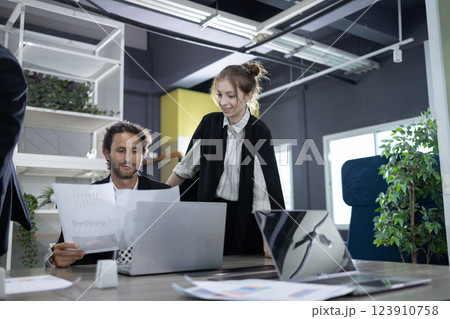 Two professionals are engaged in a discussion around a laptop in a contemporary office space. They are reviewing documents and sharing ideas while surrounded by greenery. 123910758