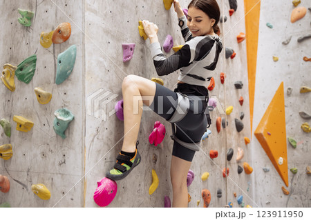 A young smiling woman on a climbing wall. A charming brunette climbs a bouldering wall. Attractive professional sport female climber having training in the gym. 123911950