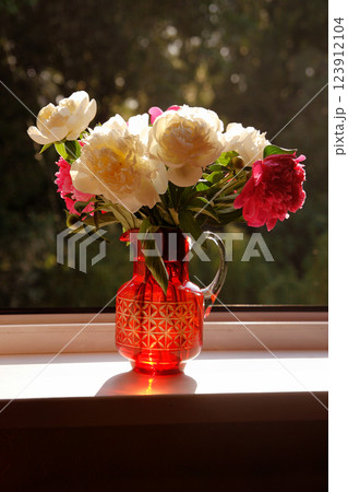 a bouquet of white and pink peonies stands in a red decanter on the window, in the rays of the morning sun a bouquet of white and pink peonies stands in a red decanter on the window, in the rays of the morning sun 123912104