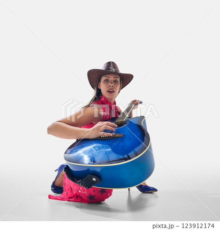 Cowgirl in vibrant outfit crouching with blue guitar, strumming with confidence and deep connection to music on white background 123912174