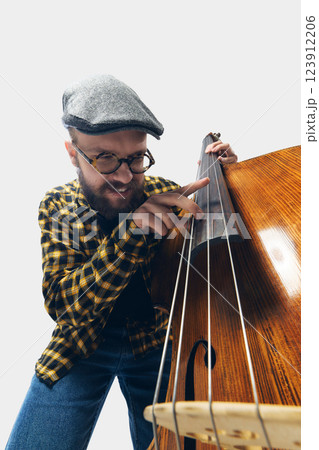 Energetic bassist, man in retro inspired outfit fully immersed into melody and performance, playing double bass against white background. 123912206