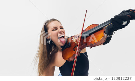 Expressive young woman violinist sticking out tongue, breaking classical norms, making passionate performance, playing violin against white background Expressive young woman violinist sticking out tongue, breaking classical norms, making passionate performance, playing violin against white background 123912227