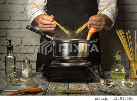 A chef in a black apron prepares pasta, holding dry spaghetti and a piece of chicken above a boiling pot. Fresh ingredients and spices decorate the kitchen countertop 123913586
