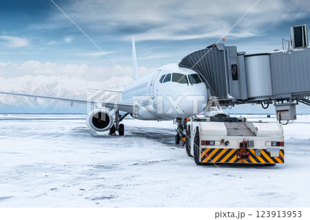 The tow tractor approaches the white passenger airplane at the aerobridge on the background of high snow covered mountains 123913953