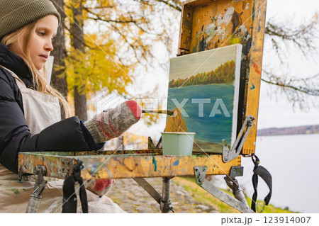 Young artist painting a landscape outdoors near a river on a cold autumn day Young artist painting a landscape outdoors near a river on a cold autumn day 123914007