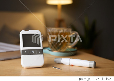 A glucometer sits on a wooden table, next to a tea cup. A blood sugar test strip and a pen are nearby, highlighting a moment of health monitoring in a comfortable setting A glucometer sits on a wooden table, next to a tea cup. A blood sugar test strip and a pen are nearby, highlighting a moment of health monitoring in a comfortable setting 123914192