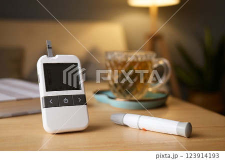A glucometer sits on a wooden table next to a cup of tea. The cozy indoor atmosphere is enhanced by soft lighting and a nearby plant, indicating a moment of self-care 123914193