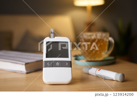 A glucometer is placed on a wooden table, showing blood sugar test results. Nearby, a cup of tea and a pen for logging data create a calming atmosphere in a cozy living space A glucometer is placed on a wooden table, showing blood sugar test results. Nearby, a cup of tea and a pen for logging data create a calming atmosphere in a cozy living space 123914200