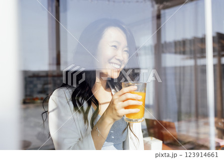 An attractive young Asian woman holds a glass of orange juice and laughs. Reflection in the window. 123914661