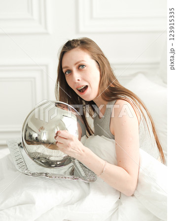 Young woman in braces and pajamas sits on bed among white sheets and opens tray. Morning breakfast in bed at hotel Young woman in braces and pajamas sits on bed among white sheets and opens tray. Morning breakfast in bed at hotel 123915335