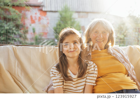 Smiling mature woman and her sweet granddaughter in backyard of house. Laughing grandmother hugs her little granddaughter. Pensioner and child are relaxing together in garden. Smiling mature woman and her sweet granddaughter in backyard of house. Laughing grandmother hugs her little granddaughter. Pensioner and child are relaxing together in garden. 123915927