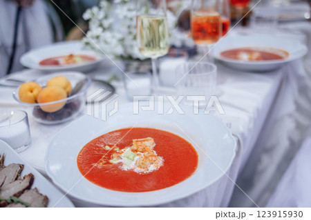 A close-up of a gazpacho plate and a covered festive table with a white tablecloth. Delicious tomato soup, glasses of wine and fruits. A close-up of a gazpacho plate and a covered festive table with a white tablecloth. Delicious tomato soup, glasses of wine and fruits. 123915930