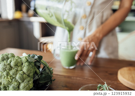 Close-up of broccoli on the kitchen table. A young woman pours a green smoothie from a blender into a glass cup. 123915932