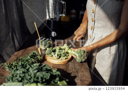 Female hands with knife, broccoli, herbs and blender. Young woman is cutting cabbage for green smoothie or salad in kitchen at home. 123915934