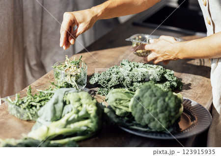 Close-up of a female hands making a green salad in the kitchen. A young woman pours pine nuts into a glass bowl with arugula, broccoli and curly cabbage leaves. Close-up of a female hands making a green salad in the kitchen. A young woman pours pine nuts into a glass bowl with arugula, broccoli and curly cabbage leaves. 123915935