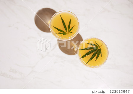 Two elegant long-stemmed glasses with green cocktails with cannabis leaves on the light marble surface of the table. A delicious healthy drink with hemp on a white background. 123915947